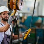 A crane operator wearing a hard hat holding a crane hook and a pendant controller in a manufacturing factory